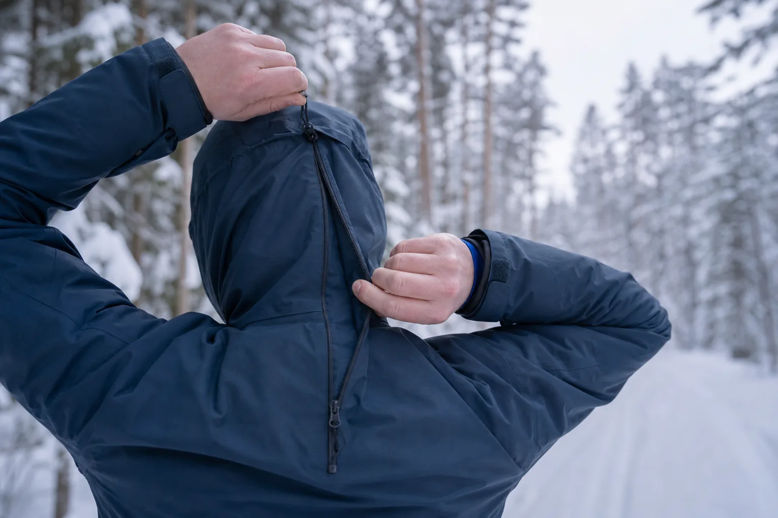Händer som justerar huva och dragkedja på en teknisk vinterjacka i snöig skog