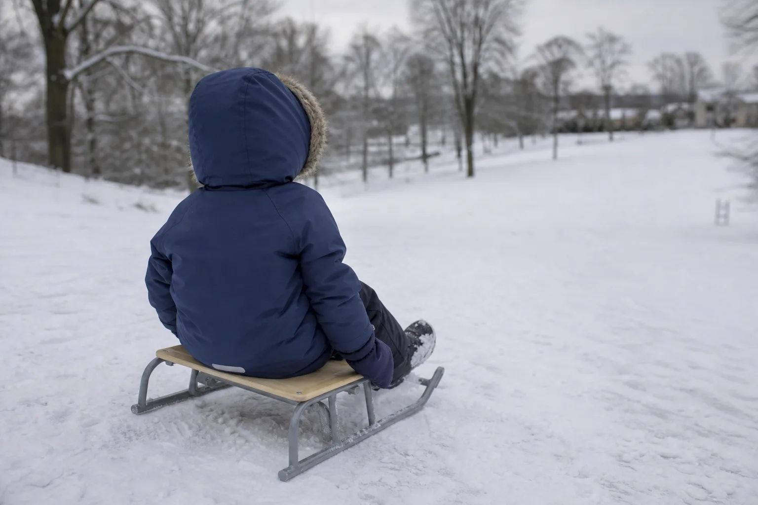 Barn sett bakifrån på pulka i en lugn snöbacke med vinterjacka och vantar